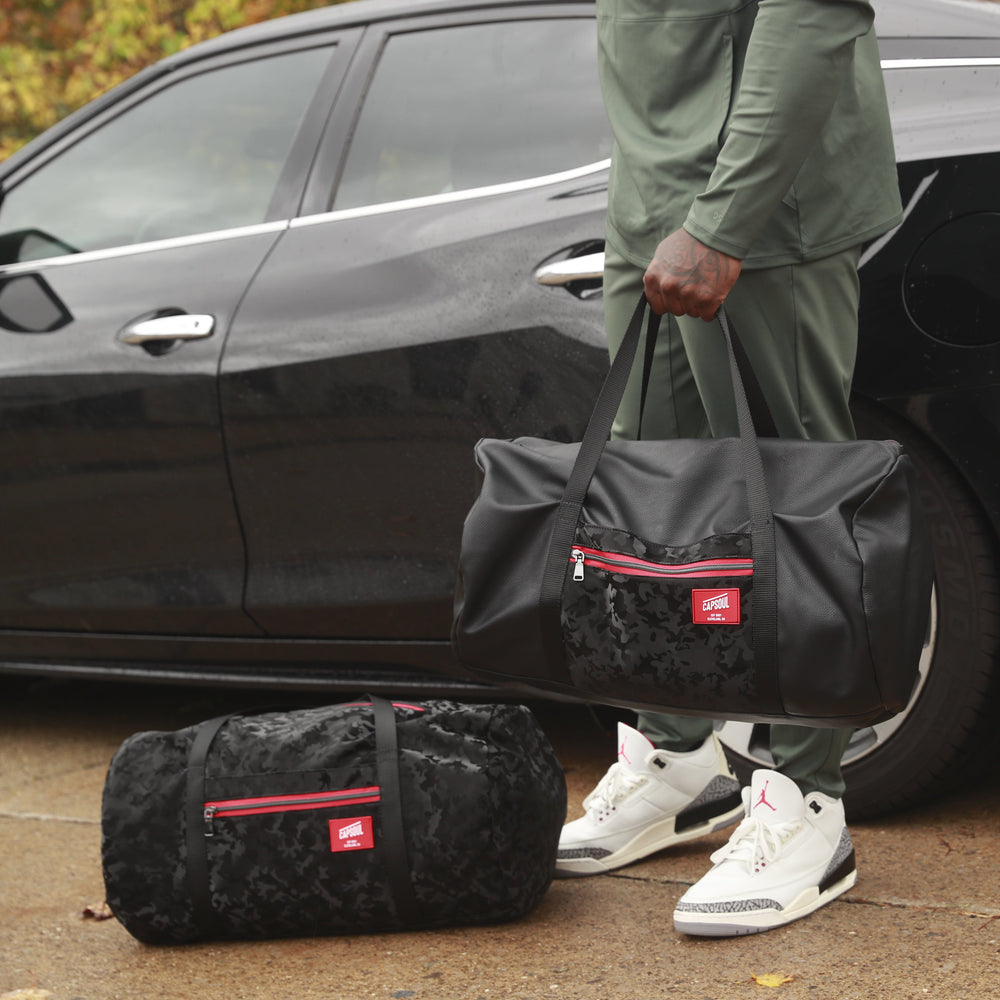 Person holding a black duffel bag with red accents next to a black car
